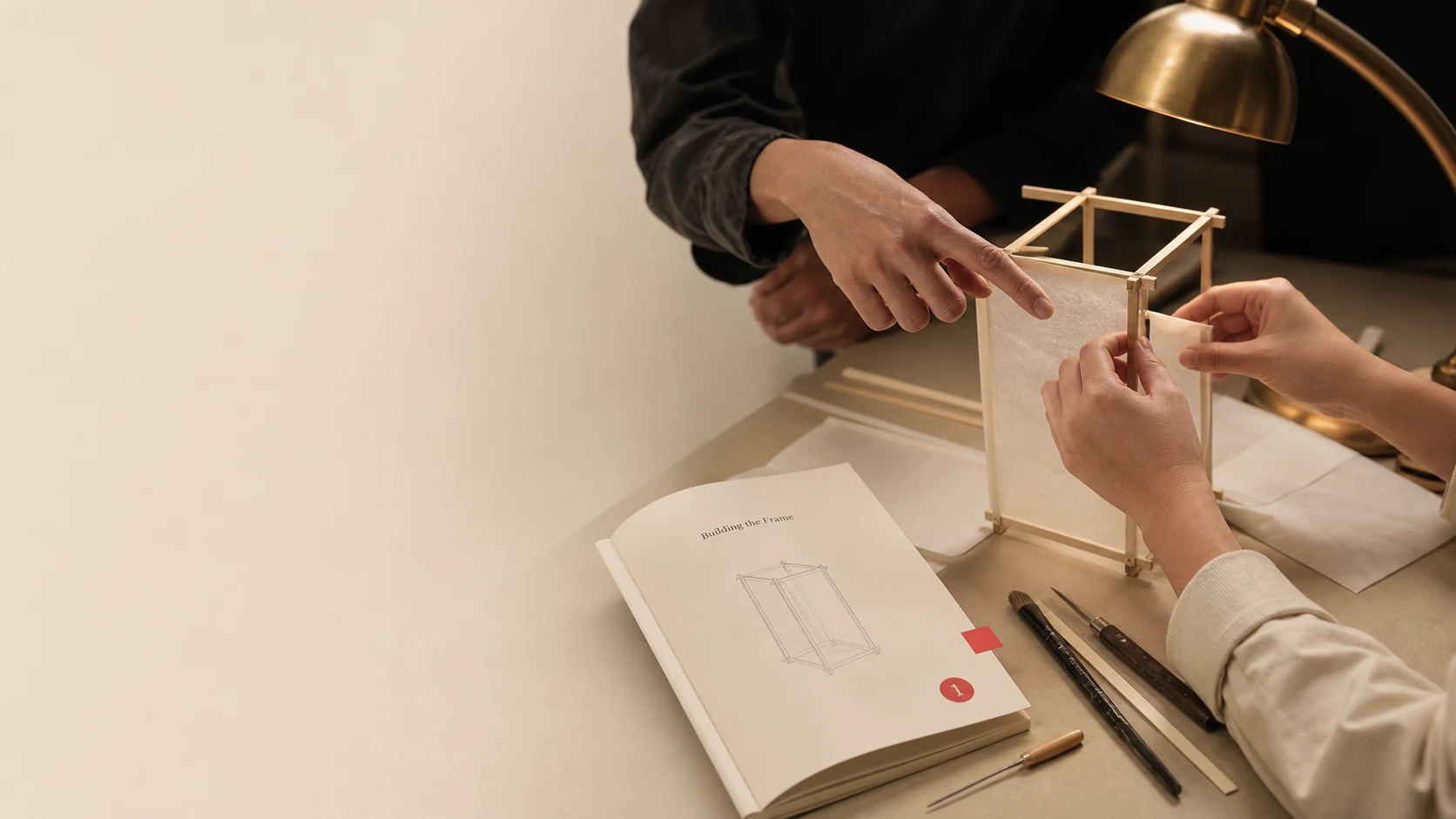 Hands assembling a lantern frame at a workbench under a brass lamp, beside a pencil drawing